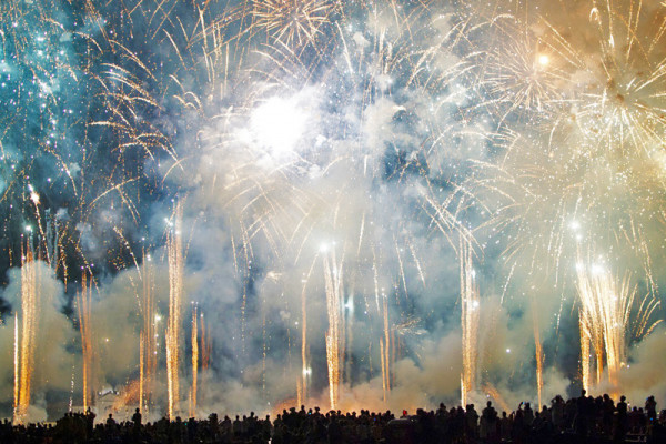 Uno spettacolo di fuochi d'artificio illuminano il cielo, con fumi e scintille, mentre una folla osserva in basso.